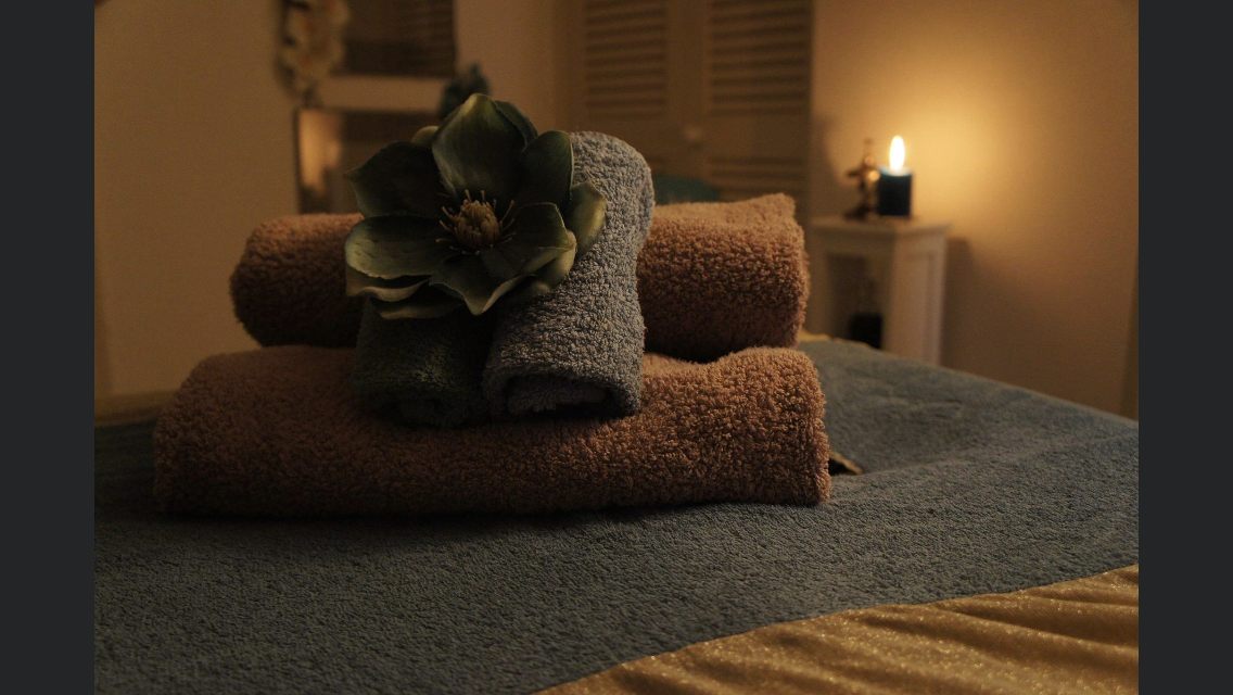 A relaxing candle casts a welcoming glow in a massage room. Soft towels are rolled up and ready for the viewer to take their pre-massage shower.